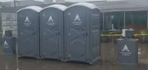 Three dark gray portable toilets and two matching handwashing stations branded with the TAP logo and website (www.TAP-NC.com) are positioned on a wet concrete surface in an industrial area
